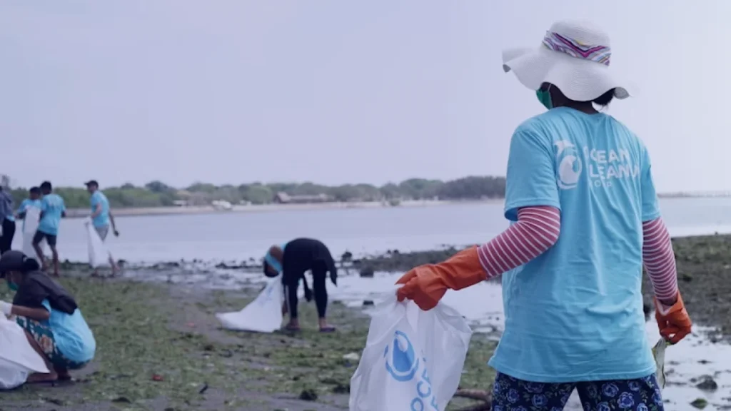 Voluntária de costas em ação de limpeza de praia, com chapéu e camiseta de organização ambiental, representando o Ano Internacional do Voluntariado 2026