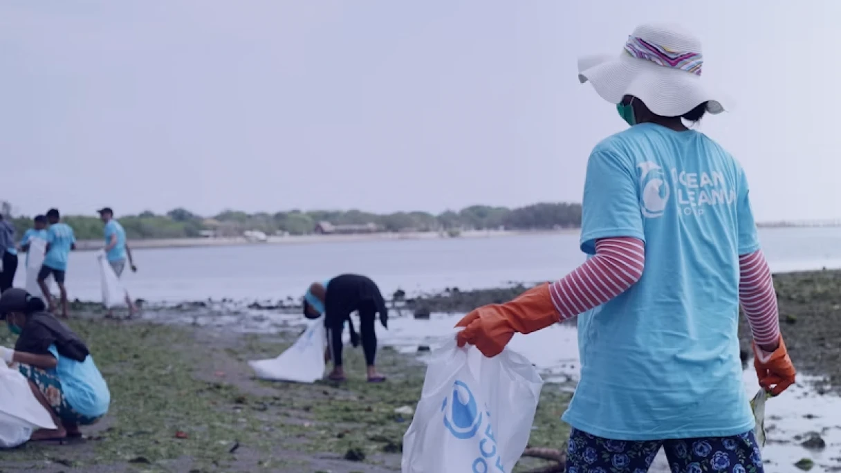 Voluntária de costas em ação de limpeza de praia, com chapéu e camiseta de organização ambiental, representando o Ano Internacional do Voluntariado 2026